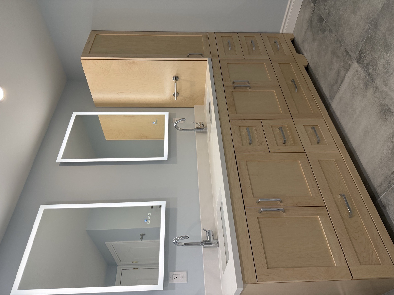 Angled view of a light-maple shaker double vanity with two illuminated LED mirrors, quartz countertop, undermount sinks, and a built-in linen tower