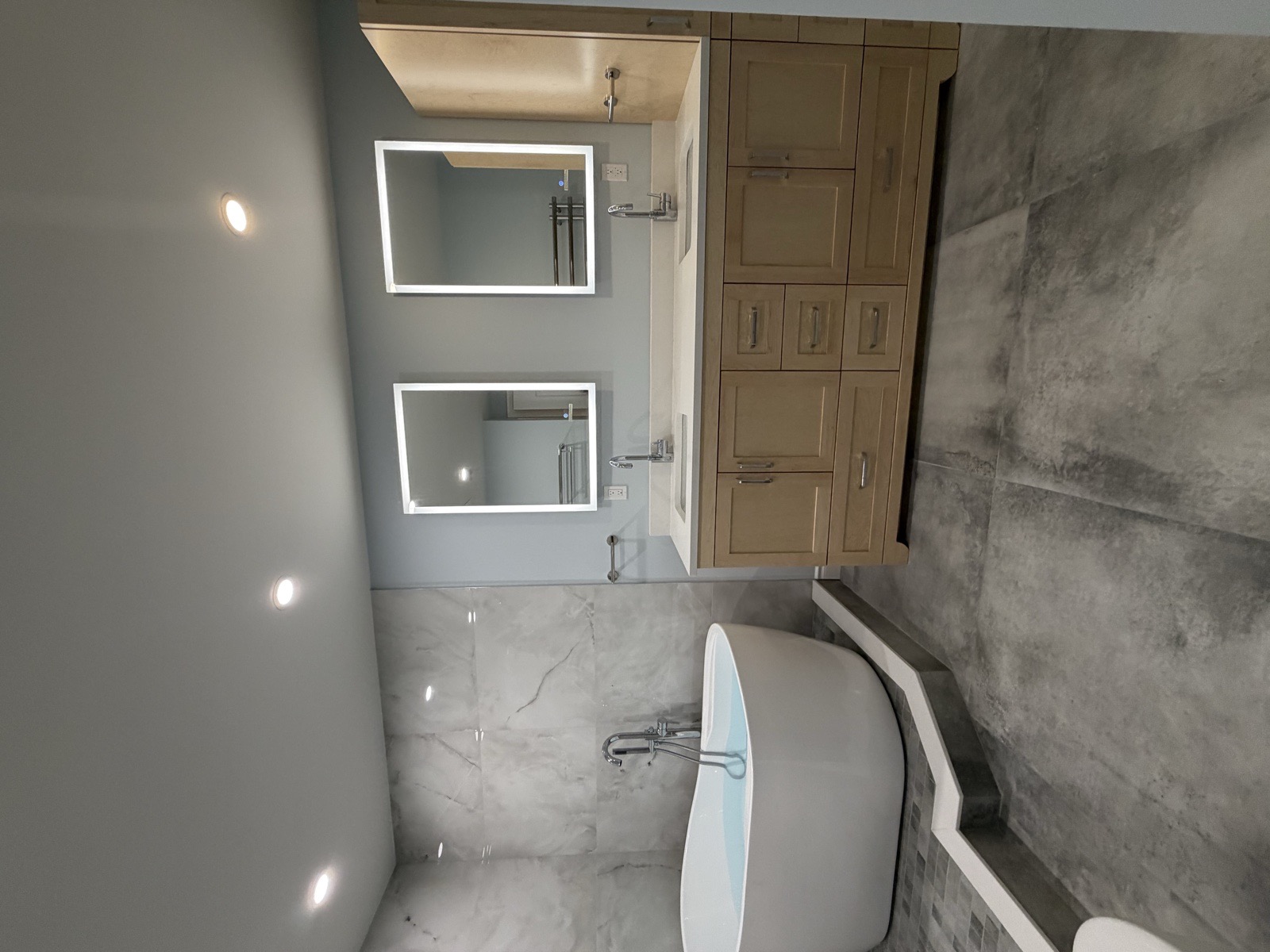 Wide view of a freestanding tub on a tiled podium across from a custom light-maple double vanity with two LED mirrors in a Mississauga condo bathroom