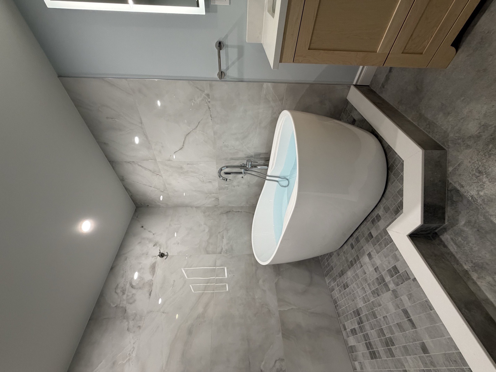 Freestanding soaking tub set on a custom-built tiled podium beside a walk-in shower, with a floor-mounted chrome tub filler and marble-look porcelain walls