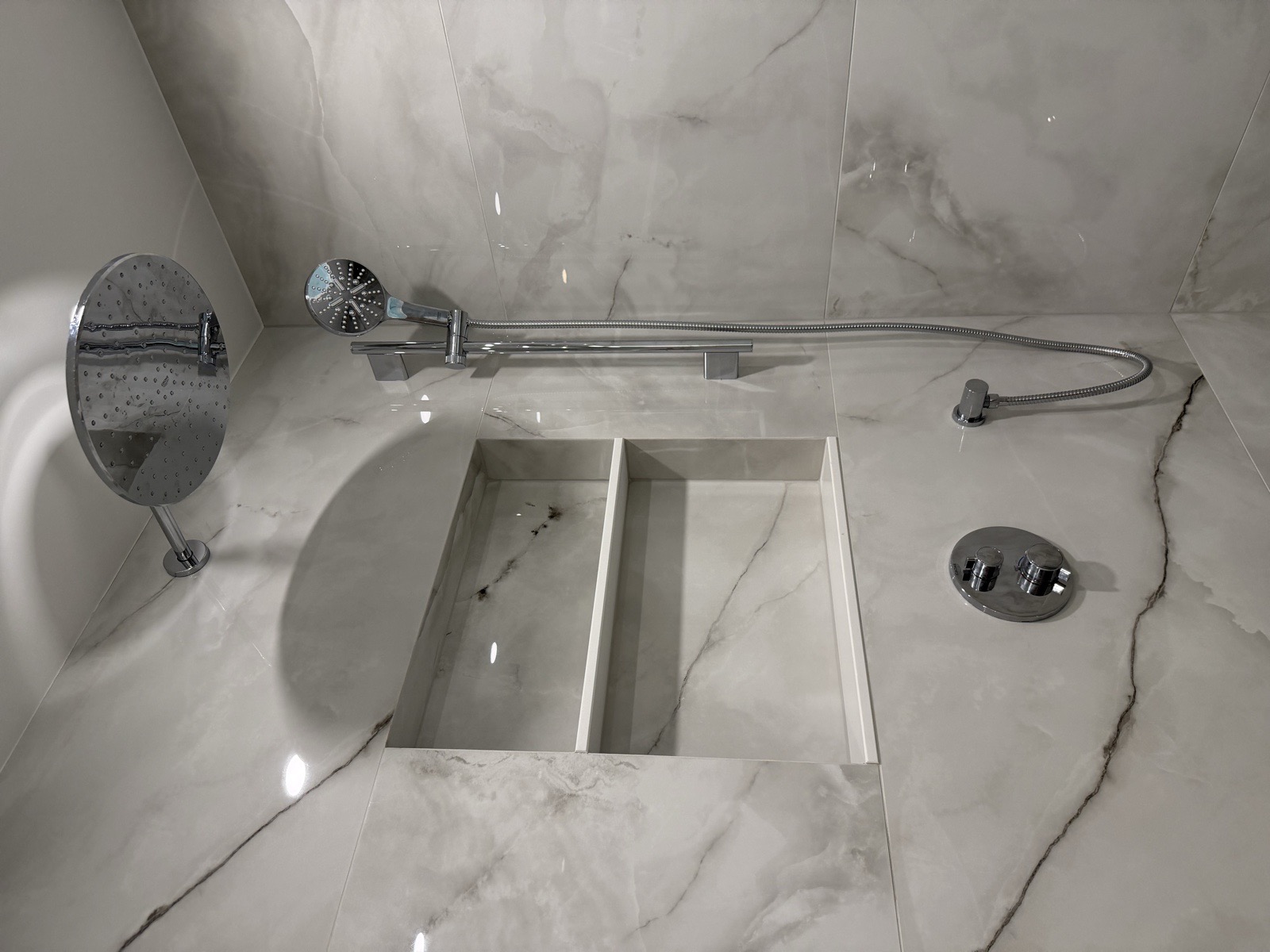 Walk-in shower interior with a round chrome rainfall head, handheld shower on a slide bar, and a custom recessed niche in marble-look tile